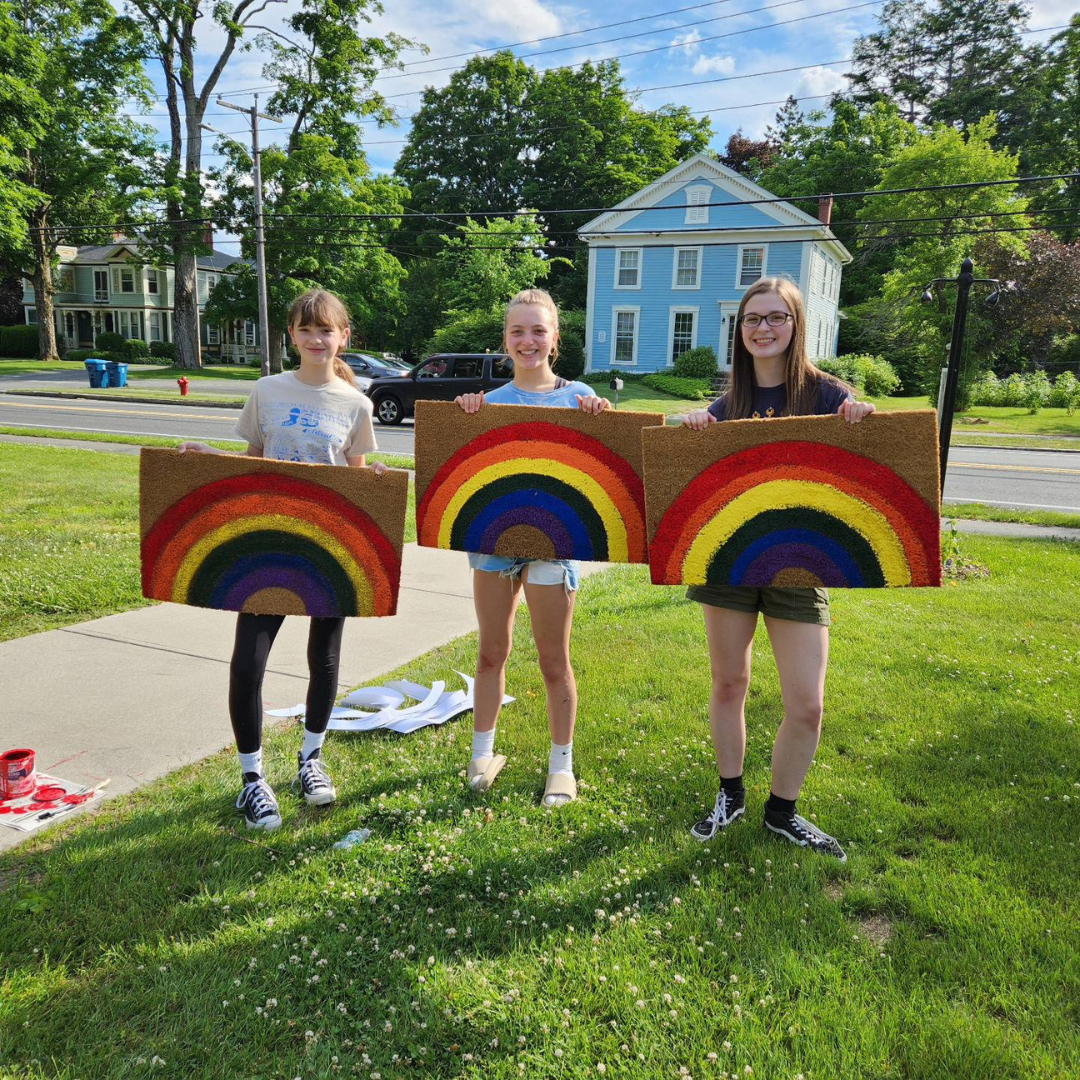 three smiling teens holding rainbow door mats they've made